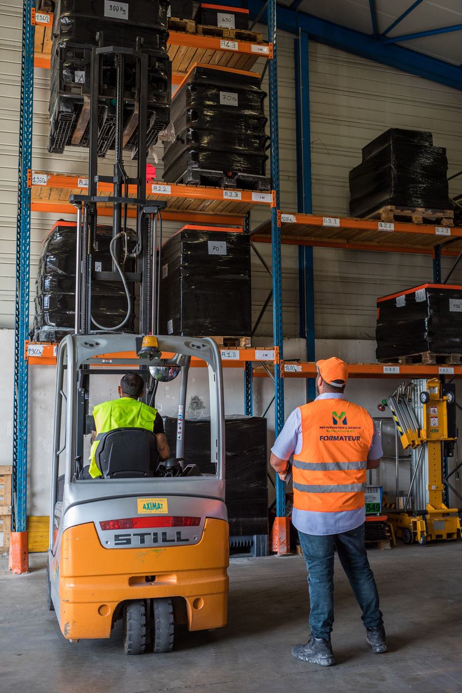Cariste manœuvrant un chariot élévateur électrique en allée de palettier, sous le contrôle du formateur